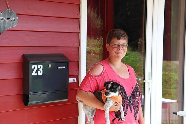 Eine junge Frau mit Hund auf dem Arm steht vor dem Eingang ihres Holzhauses